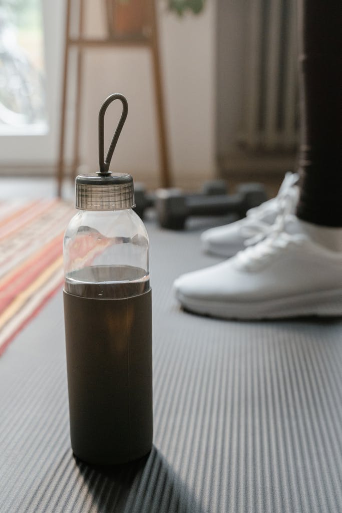 Close-up of a water bottle on a yoga mat with blurred gym equipment in a home setting.